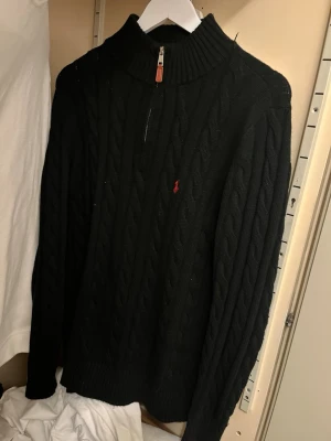 Svart kabelstickad tröja Polo Ralph Lauren - Svart kabelstickad tröja från Polo Ralph Lauren med halv dragkedja och hög krage. Tröjan har det klassiska röda broderade logot på bröstet och ribbstickade muddar. Perfekt för lager-på-lager och snygg till jeans.
