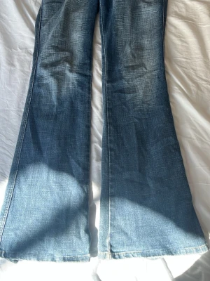 Blå bootcut jeans med slitningar - Säljer ett par blå bootcut jeans med snygga slitningar och lätt tvättad look. Jeansen har vida ben och klassisk femficksdesign. Perfekta för dig som gillar retrostil och vill ha ett par jeans med lite extra attityd.