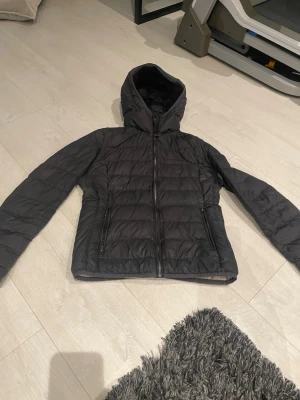 Svart parajumper jacka dam st 140-152 - Snygg svart pufferjacka med huva och dragkedja framtill. Jackan har två sidofickor med dragkedja och quiltad design som ger en sportig look. Perfekt för kalla dagar och enkel att matcha med olika outfits. Materialet är troligtvis syntetiskt och jackan är lätt vadderad.