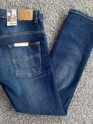 Blå jeans från Nudie Jeans - Finns i många storlekar, bara att skriva o fråga!  Säljer ett par klassiska blå jeans från Nudie Jeans med snygg tvätt och raka ben. Jeansen har fem fickor, orange sömmar och en läderpatch med logga bak. Tillverkade i slitstark denim, perfekta för en avslappnad och trendig stil.