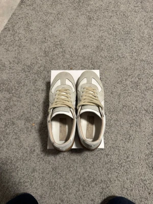 Maison Margiela Replica sneakers vita/beige - Säljer ett par Maison Margiela Replica sneakers i vitt och beige. Skorna har en klassisk låg silhuett med ovandel i mocka och läder, beigea snören och gummisula. Perfekta för dig som gillar stilrena och tidlösa sneakers. Kom med bud. 