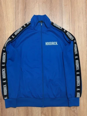 Blå zip hoodie från Hoodrich - Blå hoodie från Hoodrich med hel dragkedja och hög krage. Svarta band med vit HOODRICH-text längs ärmarna och logga på bröstet. Tillverkad i mjukt material och har ribbade muddar vid ärmslut och nederkant. Perfekt för en streetwear-look.
