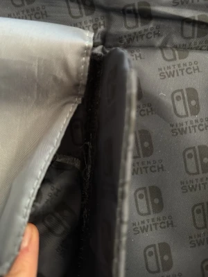 Nintendo Switch svart väska - Säljer en svart väska från Nintendo Switch med vadderad insida och flera fack för tillbehör. Väskan har dragkedja och är fodrad med grått tyg med Nintendo Switch-logga. Perfekt för att bära och skydda din Switch och tillbehör.