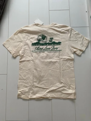 Aimé Leon Dore beige t-shirt - Snygg beige t-shirt från Aimé Leon Dore med grönt tryck på både bröstet och ryggen. Klassisk passform med rund hals och korta ärmar. Tillverkad i mjuk bomull, perfekt för en avslappnad och trendig stil.