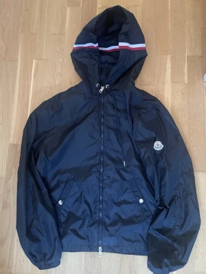 Svart vindjacka från Moncler med nfc S/M - Snygg marinblå vindjacka från Moncler med huva och dragkedja framtill. Jackan har vita och röda ränder på huvan, två fickor med tryckknappar och Moncler-logga på ärmen. Tillverkad i lätt, glansigt material som står emot vind. Kanske dene marinblå men tror de svart. Den är size 3 och de motsvarar L men dene passar mej som tar size M 