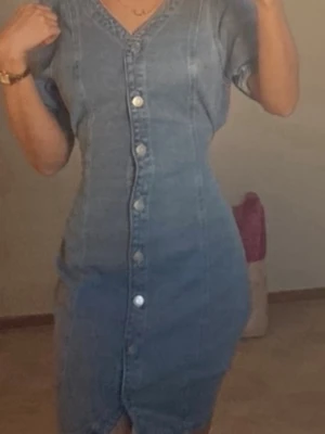 Blå jeansklänning med knappar - Snygg blå jeansklänning med kort ärm och knappar framtill. Klänningen har en v-ringad hals och markerad midja som ger en feminin siluett. Perfekt för dig som gillar en avslappnad men trendig stil.