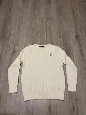 Vit kabelstickad tröja från Polo Ralph Lauren - Kabelstickad tröja i mjuk vit färg från Polo Ralph Lauren med klassisk rund halsringning och broderad logga på bröstet. Tröjan har långa ärmar och ribbade muddar vid ärmslut och nederkant. Perfekt för en stilren look.