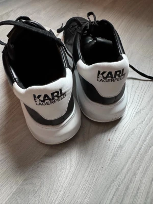 Karl Lagerfeld svarta sneakers str 44 - Snygga svarta sneakers från Karl Lagerfeld med vita detaljer och logga på hälen. Skorna har klassisk låg profil, svarta skosnören och vit sula. Tillverkade i slätt läder som ger en clean och modern look.