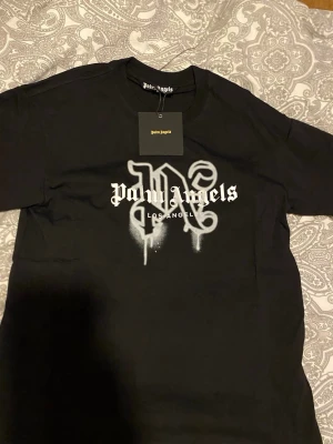 Svart Palm Angels t-shirt med tryck - Palm Angels svart t-shirt med stort vitt tryck framtill och texten 'Palm Angels Los Angeles' i en cool streetstil. Klassisk passform med rund hals och korta ärmar. Perfekt för dig som gillar trendiga och snygga plagg med attityd.