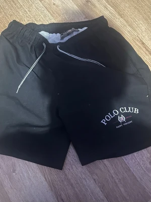 Svarta badbyxor från Polo Club - Snygga svarta badbyxor från Polo Club med vitt tryck och logga på ena benet. De har elastisk midja med vit dragsko och meshfoder på insidan för extra komfort. Perfekta för stranden eller poolen.