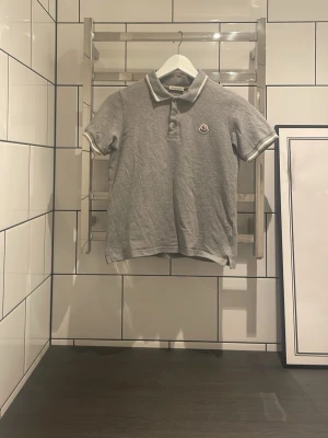 Grå pikétröja från Moncler - Klassisk grå pikétröja från Moncler med vit kant på krage och ärmslut. Tröjan har korta ärmar, tre knappar framtill och Moncler-logga på bröstet. Perfekt för en clean look. Dessvärre har tröjan tecken på användning och är lite urtvättad. Som alltid så kan jag gå ner i pris ⚜️