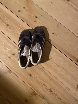Adidas Originals svarta sneakers - Säljer ett par klassiska Adidas Originals sneakers i blå mocka med vita detaljer och de ikoniska tre ränderna på sidorna. Skorna har vit sula och blå snörning. Perfekt för dig som gillar retrostil och streetwear.