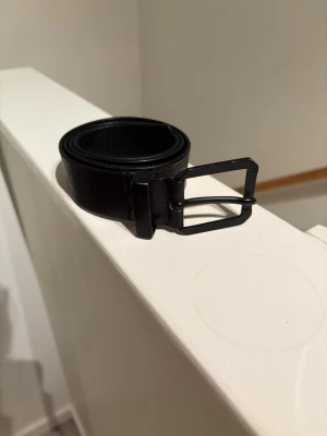 Stone Island bälte  - Stilrent svart bälte i skinn med enkel, fyrkantig metallspänne. Perfekt till jeans eller kostymbyxor för en clean och tidlös look. Bältet har flera hål för justerbar passform och en slät yta utan mönster.