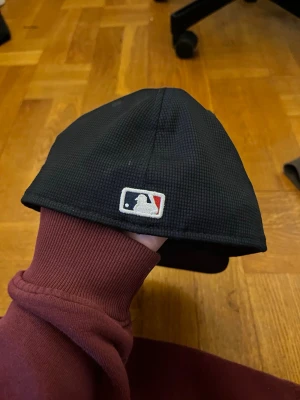 Svart MLB keps med röd logga - Säljer en svart keps från MLB med en röd och vit broderad logga framtill och klassisk MLB-logga baktill. Kepsen har böjd skärm och är tillverkad i ett strukturerat tyg som ger en sportig vibe. Perfekt för dig som gillar streetwear och baseball. pris går att diskutera 