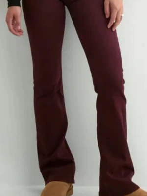 Vinröda bootcut jeans med låg midja - Snygga vinröda jeans med bootcut-modell och låg midja. Jeansen har två silverfärgade knappar, bälteshällor och klassisk femficksdesign. Perfekta för dig som vill ha en trendig och bekväm look.