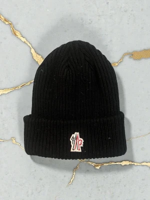 Svart ribbstickad mössa Moncler - Svart ribbstickad mössa från Moncler med uppvikt kant och broderad logotyp framtill i vitt och rött. Klassisk och stilren modell som passar perfekt till kalla dagar. Materialet ser ut att vara mjukt och värmande, troligen ull eller ullblandning.