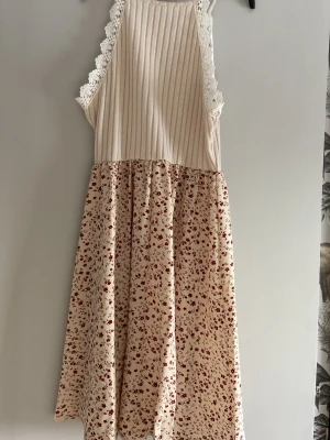 Blommig beige klänning med spets - Söt beige klänning med ribbad överdel och vita spetsdetaljer längs ärmhålen. Kjoldelen är blommig i rött och beige, vilket ger en romantisk vibe. Tillverkad i mjuk polyesterblandning med lite stretch för extra komfort.