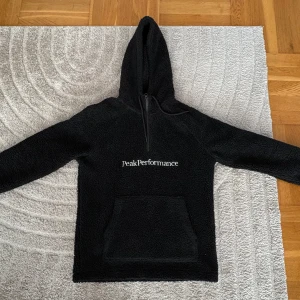Svart teddyjacka Peak Performance - Mysig svart hoodie från Peak Performance i fleece med stor magficka och dragkedja vid halsen. Hoodien har broderad logga på bröstet och en klassisk huva. Perfekt för kalla dagar när du vill vara både varm och stilren. Knappt använd.