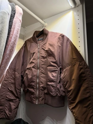 Brun oversized bomberjacka - Säljer en brun oversized bomberjacka från Weekday med breda ärmar, ribbade muddar och dragkedja framtill. Jackan har en klassisk bomberlook med hög krage och två sidofickor. Materialet är glansigt och ger en cool streetwear-vibe.