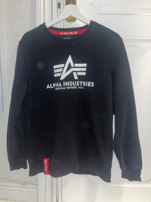 Svart sweatshirt Alpha Industries - Snygg svart sweatshirt från Alpha Industries med stort vitt logotryck på bröstet och röd detalj vid nederkanten. Klassisk rund halsringning och mjukt material som passar perfekt till streetwear-stilen.