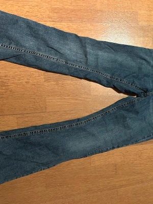 Blå bootcut jeans med fickor bak - Säljer ett par klassiska blå bootcut jeans i denim med två fickor bak som har silvriga knappar. Jeansen har en lätt utsvängd passform nedtill och normal midja. Perfekta för dig som gillar en avslappnad men ändå trendig stil.
