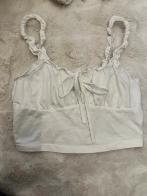 Vit linnetopp med volangband - Söt vit linnetopp med volangprydda axelband och knyt framtill. Toppen har en lätt rynkad överdel och är perfekt för varma dagar. Mjuk och bekväm känsla.