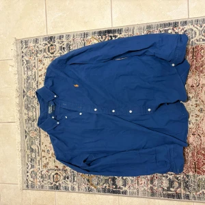 Blå skjorta från Polo Ralph Lauren - Snygg blå skjorta från Polo Ralph Lauren med klassisk button-down krage och broderad logga på bröstet. Skjortan har långa ärmar och vita knappar framtill. I princip nyskick, inga defekter. L men passar M också