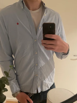 Ljusblå skjorta från Morris Stockholm - Snygg ljusblå skjorta från Morris Stockholm i storlek L. Skjortan har button-down krage, vita knappar och en liten röd brodyr på bröstet. Tillverkad i bomull med Twifold Yarn för extra kvalitet. Perfekt till jeans eller chinos.