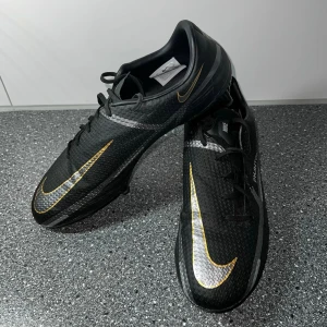 Nike Phantom GT2 fotbollsskor svart/guld - Nike Phantom GT2 fotbollsskor i svart med guldfärgade och silverfärgade detaljer. Skorna har låg profil, snörning och ovandel i strukturerat syntetmaterial för grym bollkontroll. Perfekta för dig som vill ha stilrena och funktionella skor på planen. Orginal pris 1000 kr