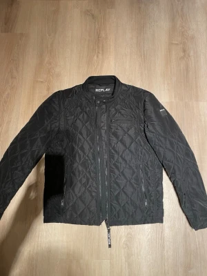 Svart quiltad dunjacka från Replay - HELT NY, Snygg svart dunjacka från Replay med quiltat mönster, ståkrage och dragkedja framtill. Jackan har tre fickor med dragkedja och Replay-logga på ärmen. Perfekt för kalla vinterdagar och har en modern, stilren look.