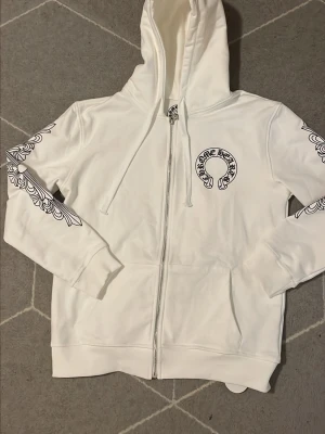 Vit Chrome Hearts zip hoodie - Säljer en vit zip hoodie från Chrome Hearts med stora svarta tryck på ryggen och bröstet samt detaljerade mönster längs ärmarna. Hoodien har dragsko i huvan och fickor framtill. Perfekt för dig som gillar streetwear och vill sticka ut.