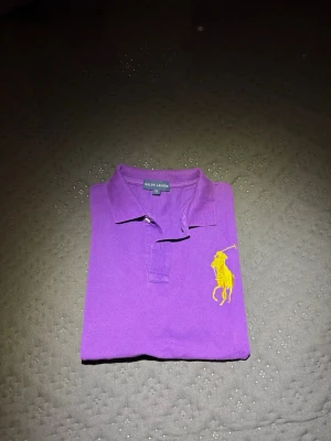 Lila pikétröja från Ralph Lauren - Säljer en lila pikétröja från Ralph Lauren med klassisk krage och knappar framtill.  Skicket är väldigt bra och har inga som helst tecken på användning. Bara att höra av dig vid eventuella frågor eller funderingar!!
