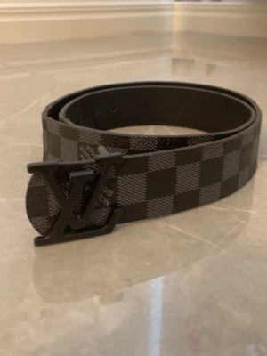 rutigt svart bälte - Snyggt svart bälte  med klassiskt rutigt mönster och stor LV-logga som spänne. Bältet är gjort i skinn och har en stilren design som passar till många outfits.