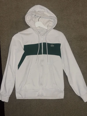 Vit hoodie med grön detalj från Lacoste - Snygg vit hoodie från Lacoste med dragkedja och en bred grön panel över bröstet. Klassisk krokodillogga på vänster sida och två fickor framtill. Tillverkad i mjukt bomullsmaterial och har justerbar huva med snören.