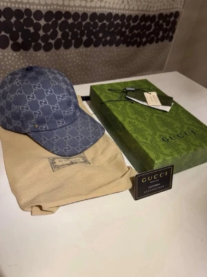 Blå GG-monogram keps från Gucci - Snygg blå keps från Gucci med det klassiska GG-monogrammönstret över hela kepsen. Kepsen har böjd skärm och justerbar rem bak. Tillverkad i denimliknande material och levereras med originalförpackning och dustbag.