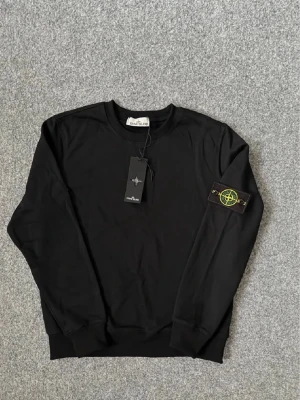 Stone Island tröja  - Säljer en svart långärmad topp från Stone Island med klassisk rund hals och ikonisk patch på ärmen. Tillverkad i mjuk bomull för skön känsla och stilren look. Perfekt för dig som gillar streetwear och vill ha något enkelt men ändå snyggt.