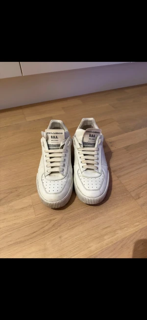 Vita sneakers från Dolce & Gabbana - Stilrena vita sneakers från Dolce & Gabbana med perforerad tå, klassisk snörning och logga på plösen. Skorna är låga och har detaljer i vitt läder samt diskreta svarta inslag på hälen. Perfekta för dig som gillar en clean och tidlös look. En skotvätt så är dom kanon 