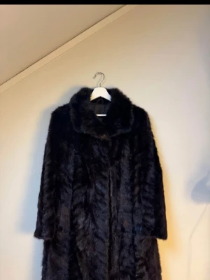Mörkbrun päls kappa med krage - Mörkbrun päls kappa med bred krage. Kappan har långa ärmar och ett lyxigt, glansigt material. Perfekt för dig som vill sticka ut och hålla dig varm under kalla dagar.         OBS ett litet hål i armhålan, men går lätt att återgärda.