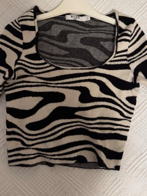 Zebramönstrad croppad topp från NA-KD - Snygg croppad topp från NA-KD med zebramönster i svart och beige. Toppen har en djup, rundad urringning och korta ärmar. Materialet är mjukt och stretchigt, perfekt för att skapa en trendig look.