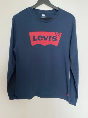 Mörkblå långärmad Levi's T-shirt  - Snygg mörkblå långärmad T-shirt från Levi's med det klassiska röda logotrycket framtill. Tillverkad i mjuk bomull och har en normal passform. Perfekt för dig som gillar enkel och stilren design med en ikonisk touch.                                                Bröstvidd: 47cm  Längd: 67cm