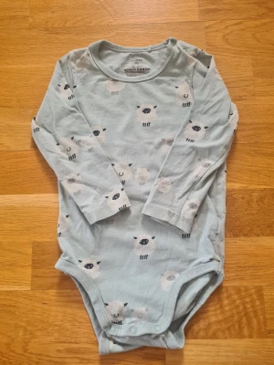 Ljusblå body med får från Lindex - Supersöt ljusblå långärmad body från Lindex  i storlek 86 med gulligt fårmönster över hela plagget. Tillverkad i mjuk bomull och har praktiska tryckknappar i grenen för enkel på- och avklädning. Finns en pytteliten fläck se sista bilden. 