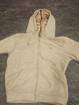 Grå zip hoodie från Burberry - Snygg grå hoodie med dragkedja från Burberry. Klassisk huva med det ikoniska rutmönstret på insidan, dragsko och två stora fickor framtill. Tillverkad i mjuk bomullsblandning, perfekt för en chill och stilren look.