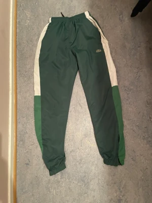 Gröna mjukisbyxor från Lacoste - Mörkgröna mjukisbyxor från Lacoste med vita sidopaneler och klassisk krokodillogga på vänster ben. Byxorna har resår i midjan och vid bensluten samt sidofickor. Perfekta för en avslappnad och sportig stil.