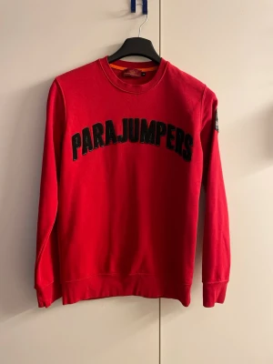 Röd sweatshirt från Parajumpers - Röd sweatshirt från Parajumpers med svart broderad logga över bröstet och patch på ärmen. Klassisk rund halsringning och ribbade muddar vid ärmslut och nederkant. Perfekt för en avslappnad och trendig stil.