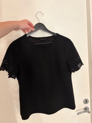 Svart topp med spetsdetaljer - Stilren svart topp med rund hals och korta ärmar. Ärmarna har snygga spetsdetaljer med utskurna mönster som ger en unik touch. Perfekt att matcha med både jeans och kjol för en trendig look.vet ej märket då jag köpt den på vintage butik
