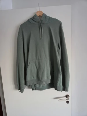 Grön hoodie med magficka - En stilren grön hoodie med huva och dragsnören. Hoodien har en stor magficka framtill och ribbade muddar vid ärmslut och nederkant. Perfekt för en avslappnad look och enkel att matcha med jeans eller joggers. Materialet känns mjukt och bekvämt.