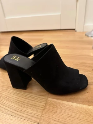 Svarta slip-in sandaler från XTI - Stilrena svarta slip-in sandaler från XTI med öppen tå och bred klack. Ovandelen är i mockaimitation och sulan är svart med diskret logga. Perfekta för dig som gillar chunky klackar och minimalistisk design.