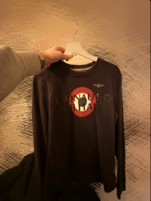 Svart långärmad tröja Aeronautica - Svart långärmad tröja från Aeronautica Militare med rund halsringning och tryckt logga samt emblem framtill i rött, vitt och svart. Perfekt för en stilren och avslappnad look.