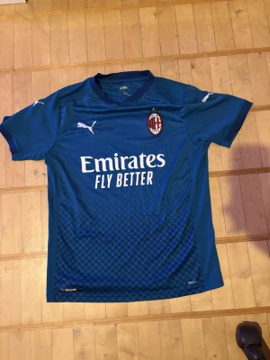 Blå AC Milan fotbollströja Puma - Säljer en blå AC Milan fotbollströja från Puma med klubbmärke och sponsortryck på bröstet. Tröjan har korta ärmar, V-ringning och är tillverkad i ett lätt och ventilerande funktionsmaterial. Perfekt för träning eller match!