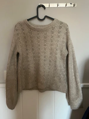 Beige stickad tröja med hålmönster - Säljer en beige stickad tröja med rund hals och snyggt hålmönster över hela plagget. Tröjan har långa ärmar och ribbade muddar vid ärmslut och nederkant. Perfekt för lager på lager och riktigt mysig till hösten.
